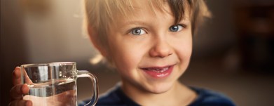 Closeup portrait of a little boy holding a glass of water. The boy is wearning blue blouse and smiling to the camera. The boy is aged 5 and is backlit by the morning sun from the window behind him Snohomish WA